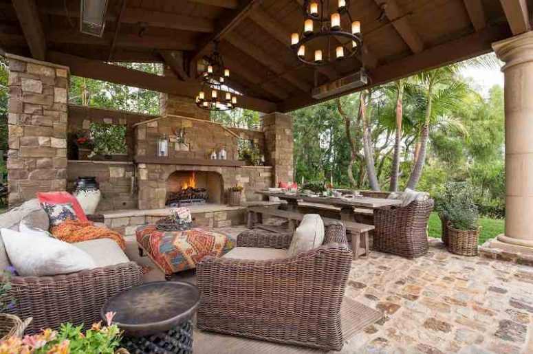 a welcoming indoor-outdoor Mediterranean living room with a fireplace in a stone wall, wicker seating furniture, a wooden table and benches and some greenery