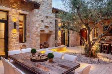 a welcoming outdoor Mediterranean space with a living tree, a wooden table, white chairs, a non-working fireplace with some lanterns