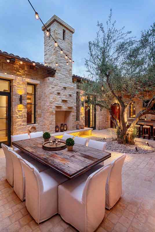 a welcoming outdoor Mediterranean space with a living tree, a wooden table, white chairs, a non-working fireplace with some lanterns