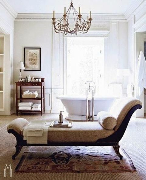 an airy vintage bathroom with a bathtub opposite the window, a dark-stained storage unit, a daybed with neutral upholstery and a pritned rug