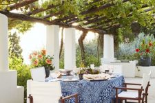 an exquisite Mediterranean patio with greenery over the space, a table with blue tablecloth and dark chairs with white upholstery and blooms