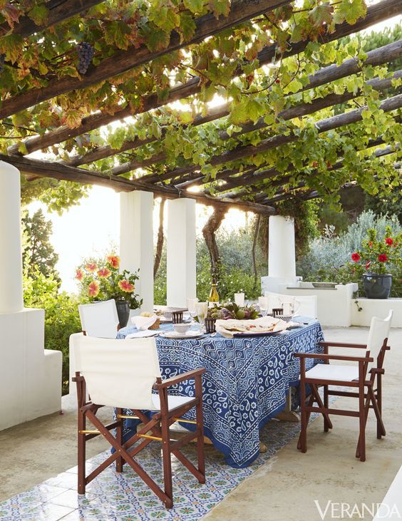 an exquisite Mediterranean patio with greenery over the space, a table with blue tablecloth and dark chairs with white upholstery and blooms