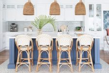 a beach kitchen with white cabinetry, a navy kitchen island, aqua green tiles on the backsplash rattan chairs, woven lamps and greenery