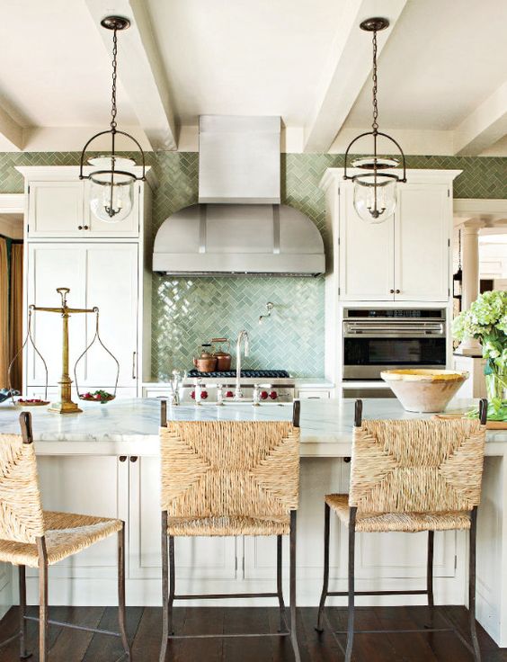 a beautiful coastal kitchen with white vintage cabinets, a large kitchen island with wicker chairs, glass pendant lamps and a green tile backsplash
