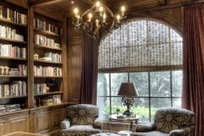 a beautiful home library fully clad with wood, with a rich-stained coffered ceiling and a vintage chandelier, printed furniture and a leathe rpouf