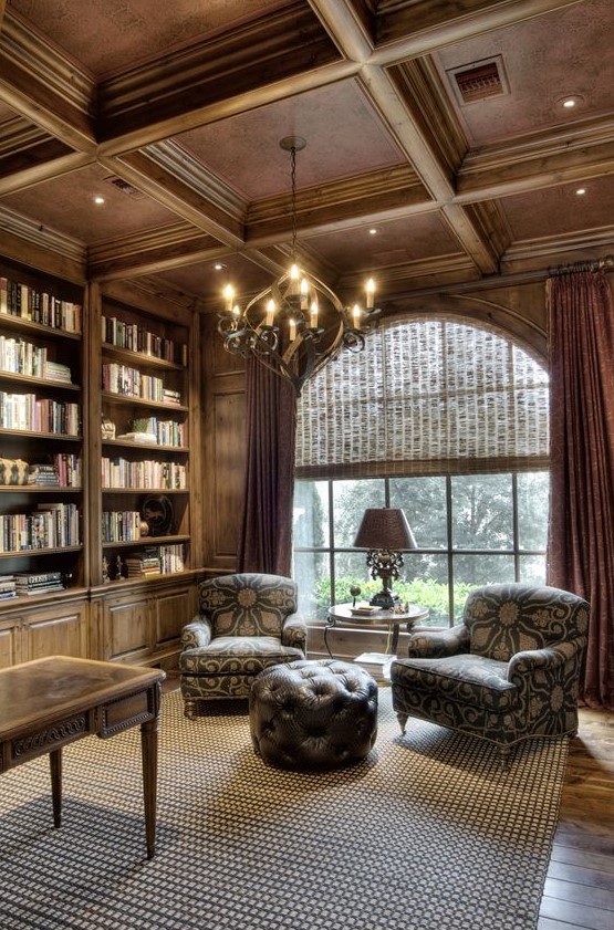 a beautiful home library fully clad with wood, with a rich-stained coffered ceiling and a vintage chandelier, printed furniture and a leathe rpouf