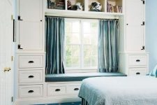 a blue and white bedroom with blue walls and a navy rug, a bed with blue bedding, a neutral storage unit with drawers and blue curtains and an upholstered bench