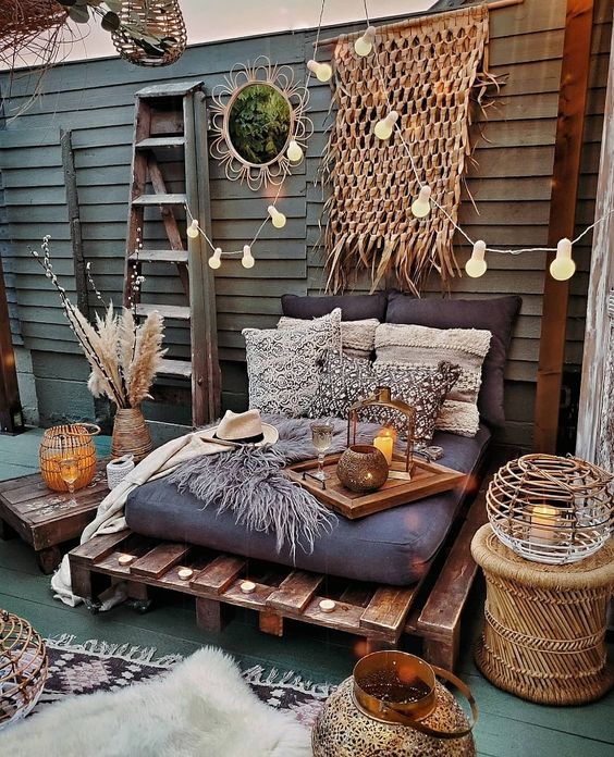 a boho outdoor bedroom with a pallet bed and black upholstery, lights, a woven hanging, candle lanterns and pampas grass
