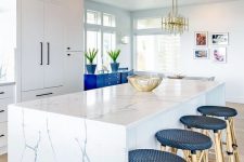 a bold and chic beach kitchen with white cabinets, a white marble kitchen island a bold blue cabinet and navy rattan stools
