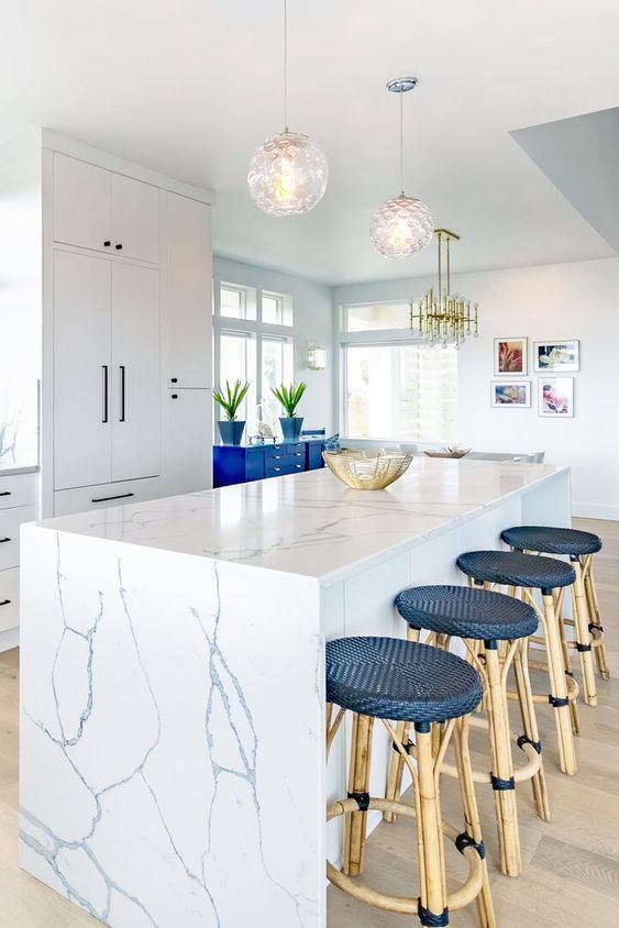 a bold and chic beach kitchen with white cabinets, a white marble kitchen island a bold blue cabinet and navy rattan stools