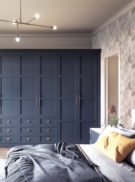 a bold bedroom with floral wallpaper, a neutral bed with blue bedding and yellow pillows, a navy wardrobe with paneled doors and drawers and a chic chandelier