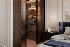 a chic bedroom with super elegant decor with a navy upholstered bed, built-in lights, a walk-in closet with glass sliding doors that add interest to the room