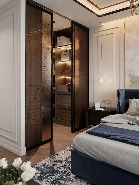 a chic bedroom with super elegant decor with a navy upholstered bed, built in lights, a walk in closet with glass sliding doors that add interest to the room