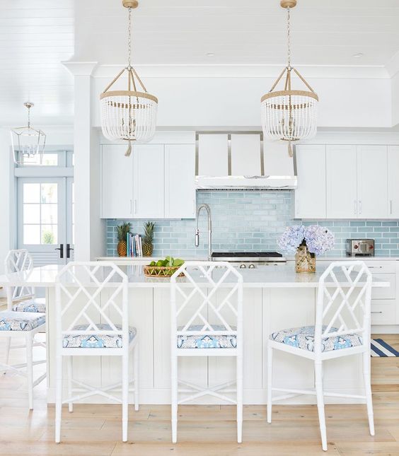 a coastal kitchen with white cabients, a large kitchen island, chic beaded chandeliers, white chairs with printed upholstery