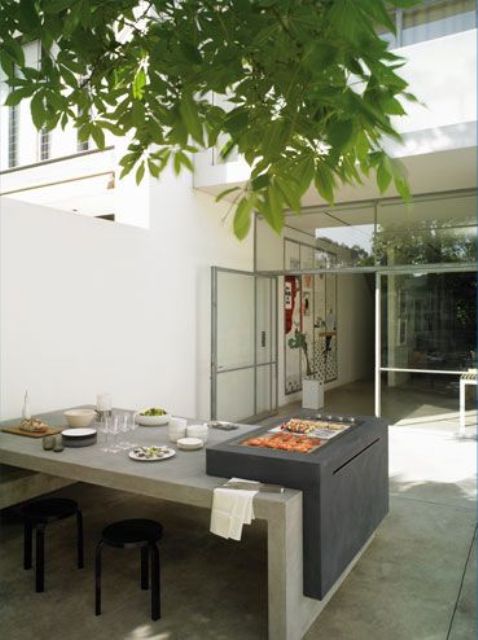 a contemporary outdoor dining space with a concrete table and an additional mini grill on top plus greenery over the table