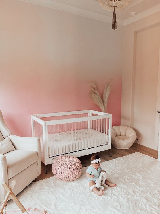 a cozy nursery with an ombre pink accent wall, creamy furniture, creamy textiles and a pink chunky knit pouf is wow