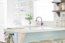 a dreamy coastal kitchen with white open shelving, an aqua kitchen island, matching pendant lamps and tableware, vintage stools and black fixtures