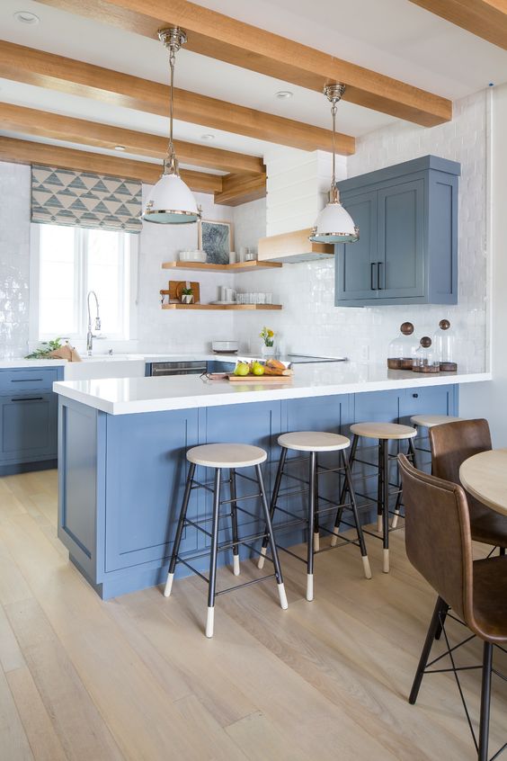 a fab coastal kitchen with blue cabinets, white stone countertops and glossy tiles, light stained shelves and wooden beams