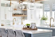 a fab coastal kitchen with white shaker style cabinets and a light blue kitchen island, glass pendant lamps and grey woven stools plus gold fixtures