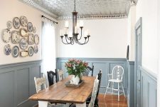 a farmhouse dining space wiht a tin tile ceiling, grey paneling, a white dining table with a stained tabletop and a gallery wall of vintage plates