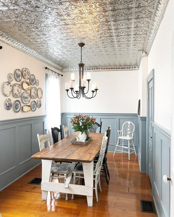 a farmhouse dining space wiht a tin tile ceiling, grey paneling, a white dining table with a stained tabletop and a gallery wall of vintage plates