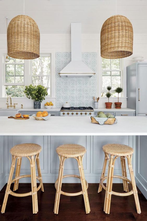 a gorgeous coastal kitchen with light blue cabinets, a light blue kitchen island, rattan stools and woven pendant lamps, potted greenery