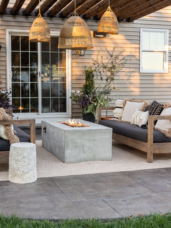 a modern outdoor space with wooden sofas with lots of pillows, a concrete fireplace, woven pendant lamps and a cocnrete stool