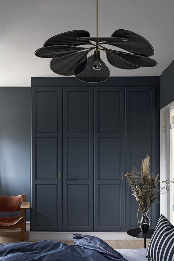 a moody bedroom with graphite grey walls and a graphite grey wardrobe with paneling, a black petal pendant lamp and a bed with a navy and white bedding