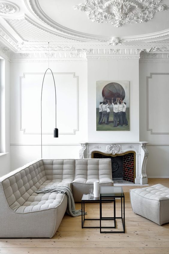 a neutral French living room with a stucco ceiling and paneled walls, a vintage fireplace, a grey modern sofa and a pouf and a floor lamp