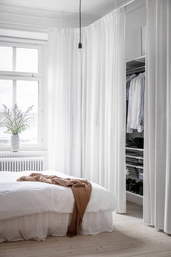 a neutral bedroom with a bed with neutral bedding, a closet with hangers and white curtains, a pendant bulb and some greenery