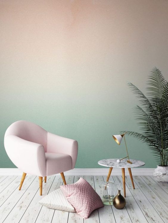 a pastel space with a blush to green gradient accent wall, a blush chair and some pillows, a potted plant and a round table