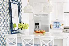 a preppy beach kitchen with white shaker cabinets, a kitchen island with white and blue stools, white woven pendant lamps and an accent wall