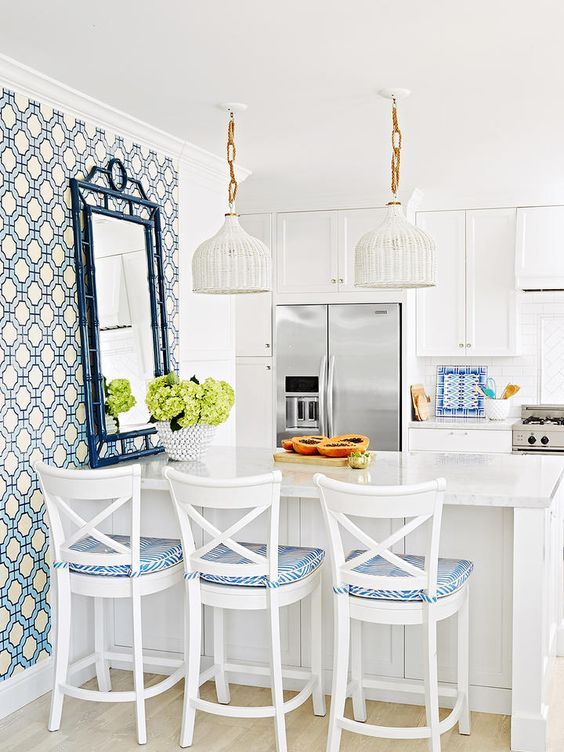 a preppy beach kitchen with white shaker cabinets, a kitchen island with white and blue stools, white woven pendant lamps and an accent wall