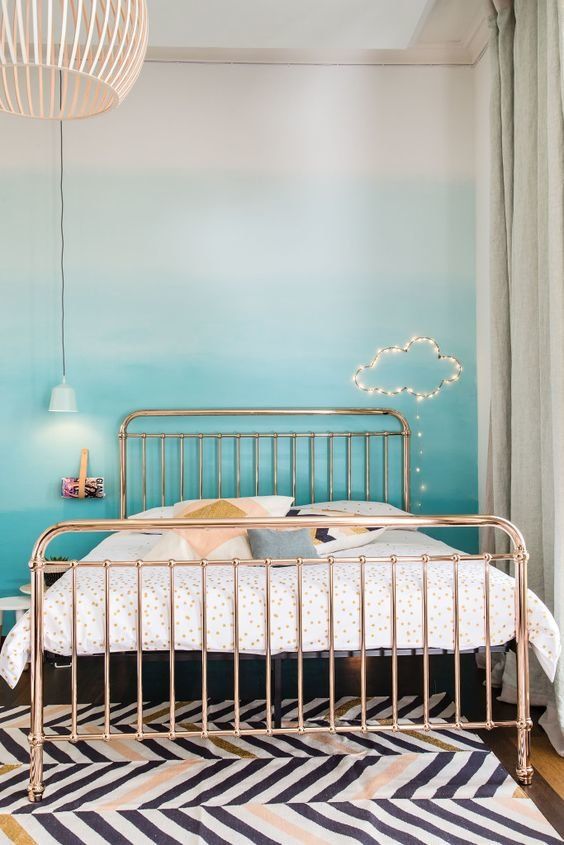 a pretty bedroom with an ombre turquoise wall, a metal bed with printed bedding, a printed rug, a cloud of string lights and pendant lamps