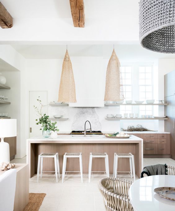 a serene coastal kitchen with stained cabinets, a large wooden kitchen island, statement wicker lamps, wooden beams and wicker chairs