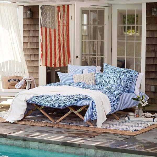 a simple outdoor bedroom next to the pool, with a bed with blue bedding, a white chair and layered rugs