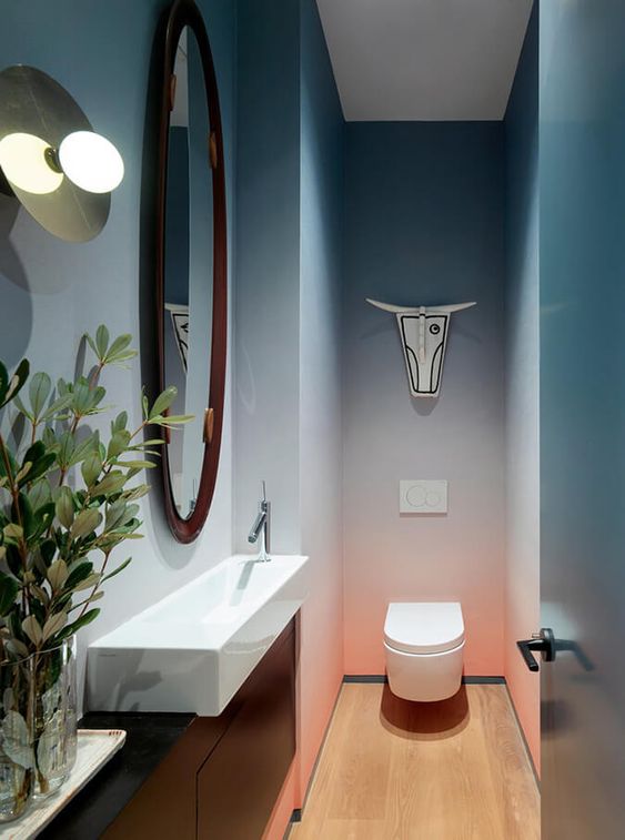 a small yet chic powder room with gradient blue to coral walls, a small sink, a toilet and a mirror plus some greenery