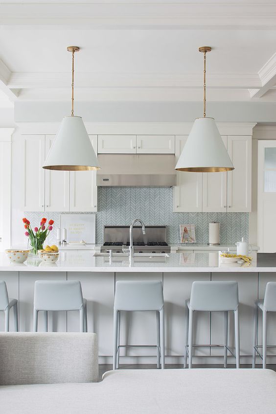 a sophisticated coastal kitchen with white shaker cabinets, a light blue herringbone backsplash, a large kitchen island, light blue stools and chic lamps