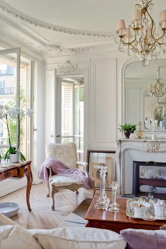 a super refined vintage French living room with a non working fireplace, molding on the walls and ceiling, a blush chandelier and sophisticated furniture