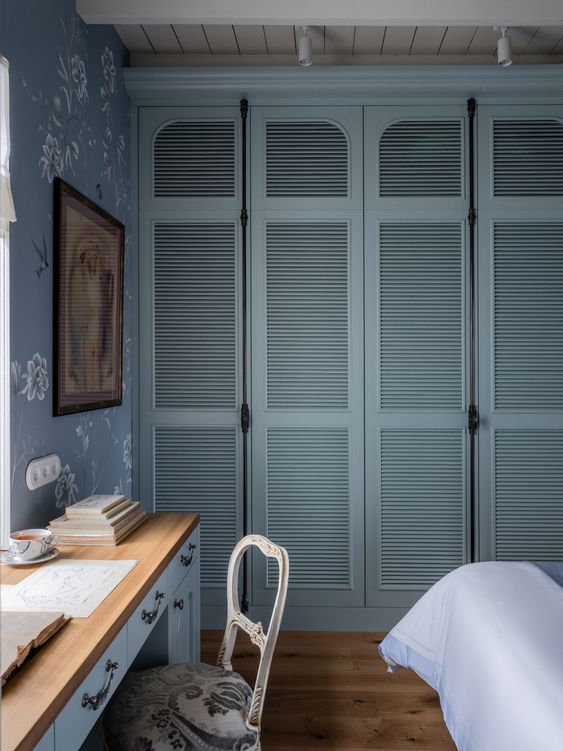 a vintage bedroom with a blue and white printed wallpaper wall, a blue desk and a vintage chair, a wall to wall wardrobe with blue shutter doors, a bed with bedding