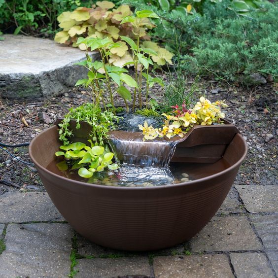 an aqua garden of a large bowl with a pond with pebbles and plants, a fountain and blooms and greenery