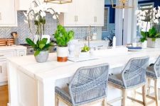 an elegant and stylish beach kitchen with white shaker cabinets, a blue herringbone backsplash, grey woven stools and brass pendant lamps
