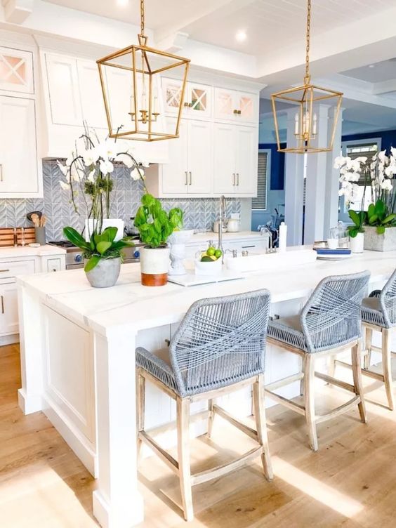 an elegant and stylish beach kitchen with white shaker cabinets, a blue herringbone backsplash, grey woven stools and brass pendant lamps