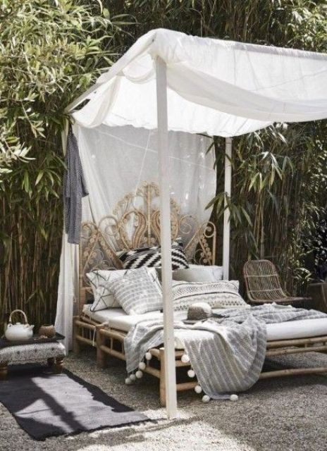 an outdoor white boho bedroom with a refined rattan and bamboo bed, neutral bedding and a canopy, a rattan chair and a low nightstand