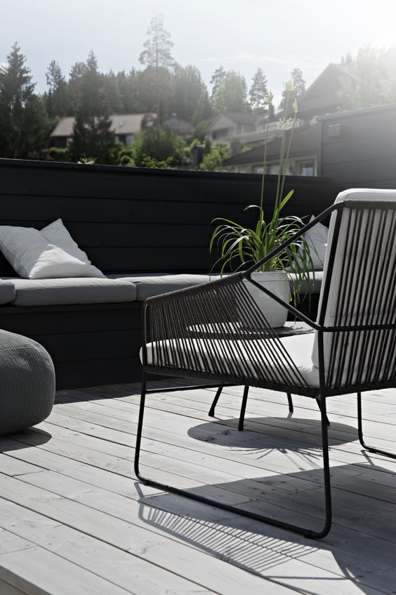 a Nordic terrace with a grey deck, a black built-in bench, a black chair with white upholstery and greenery