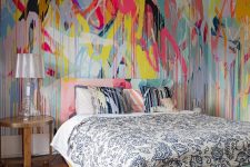 a bold bedroom with colorful graffiti all over the walls, a colorful bed with printed bedding, a nightstand and a glossy table lamp