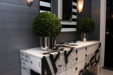 a catchy and chic entryway with grey wallpaper walls, a black and white graffiti credenza, a mirror in a large black and white frame and greenery