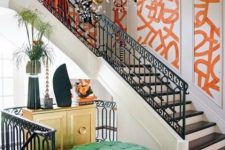 a colorful entryway with orange graffiti panels, a gilded credenza, a green tufted ottoman and a chic gold and crystal chandelier