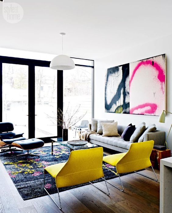 a colorful living room with a grey sofa, colorful pillows, a black chair, yellow chairs, a bold rug and a large graffiti artwork