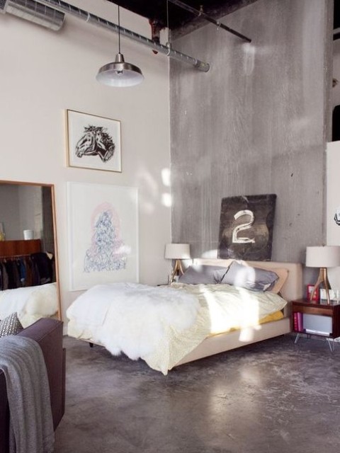 a contemporary meets industrial bedroom with a concrete wall and floor, an upholstered bed, a large mirror, pendant lamps and exposed pipes